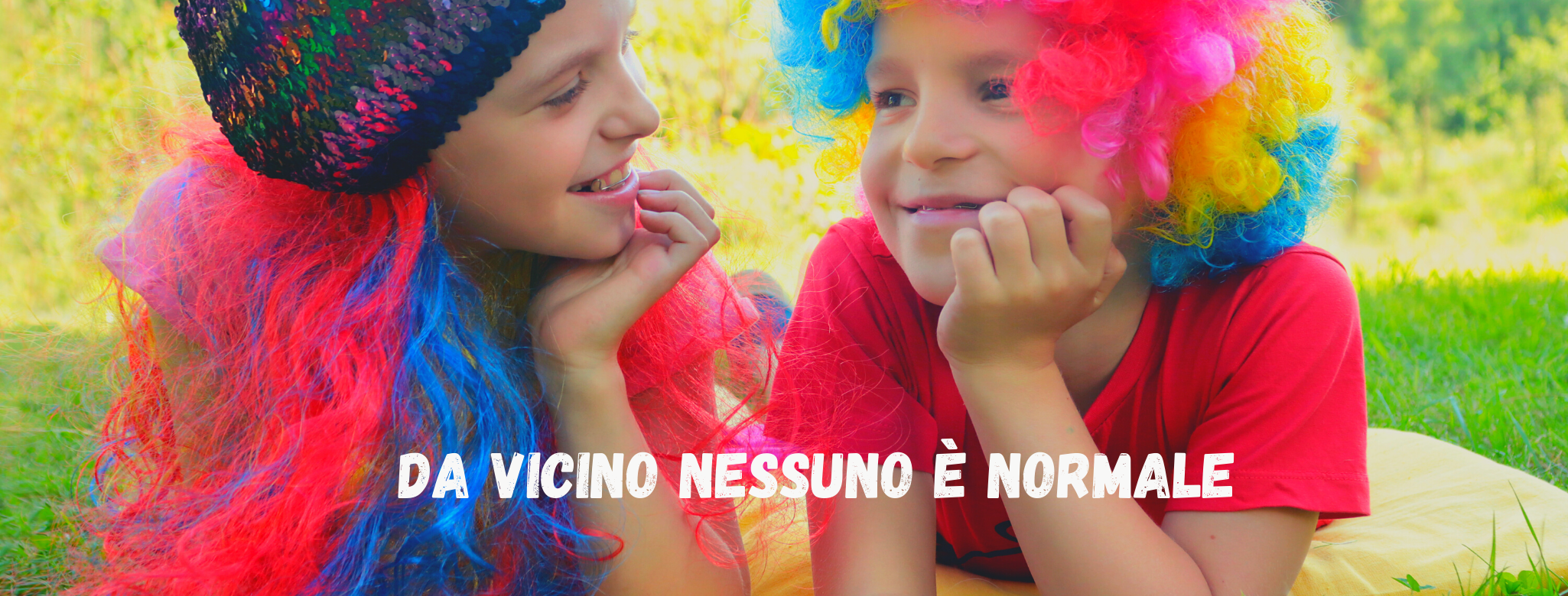 Immagine di bambino e bambina con parrucche con colori sgargianti, si guardano sorridendo: scritta "da vicino nessuno è normale"